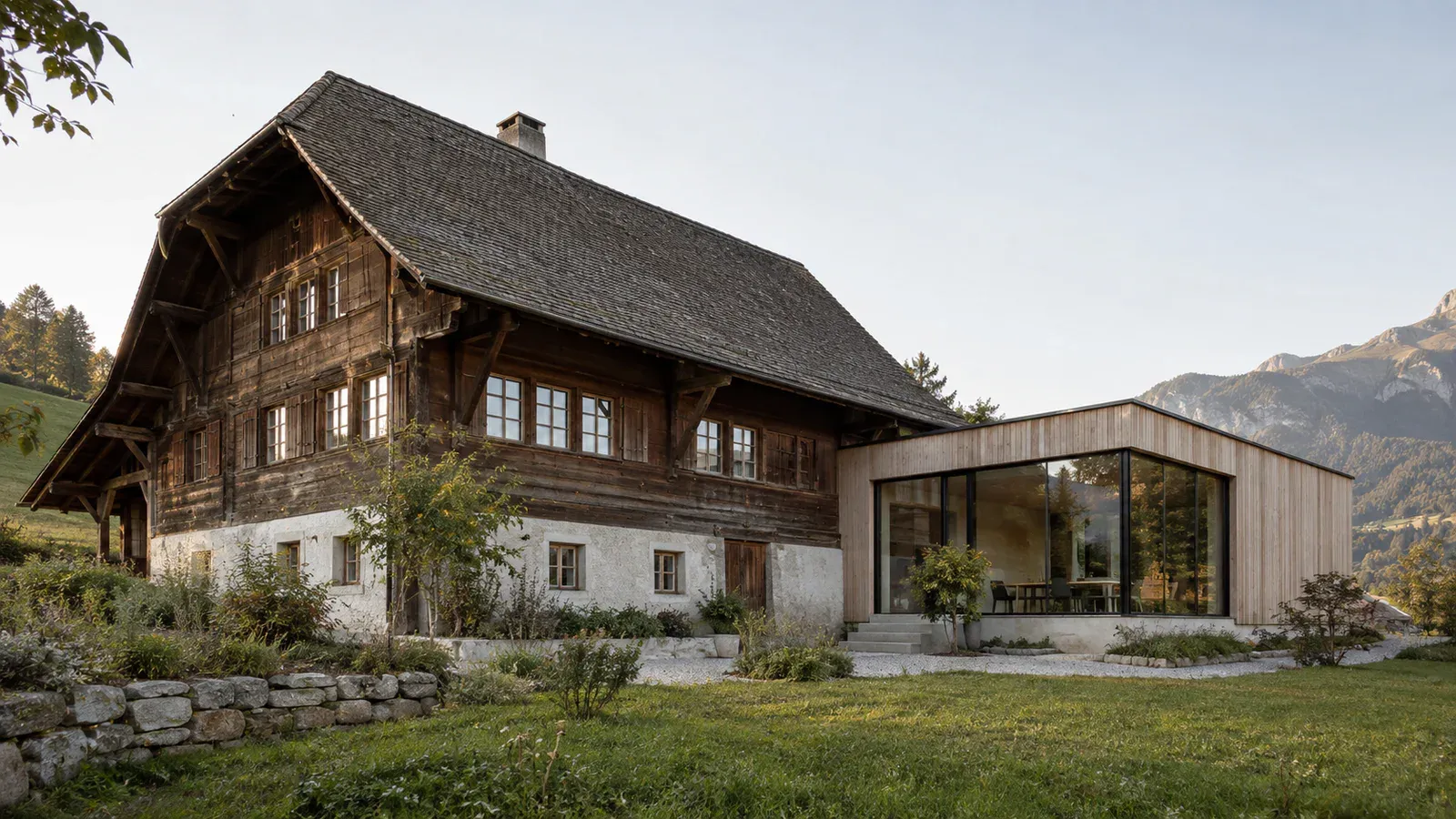 Bauernhaus aus Holz und verputztem Mauerwerk neben einer eingeschossigen modernen Holzerweiterung mit grosser Verglasung.